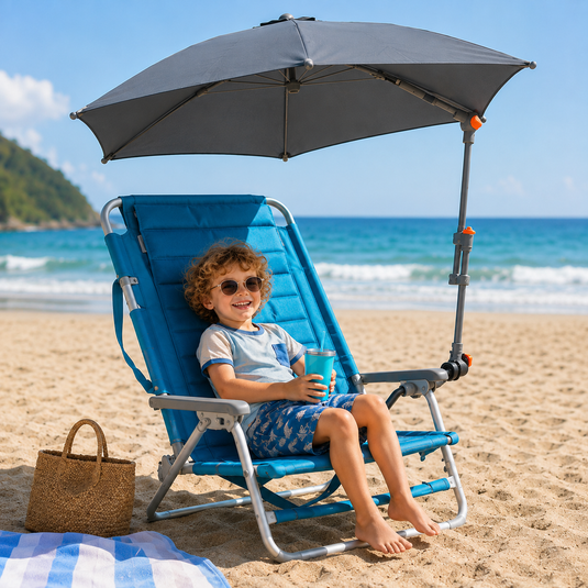 A child sits in a blue beach lounge chair with a gray sun umbrella on a sandy beach. The lounge chair has a silver metal frame, and there is a straw tote bag and a blue-and-white striped blanket nearby.